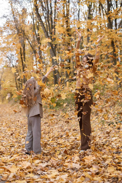 Young woman and little girl in autumn forest. Woman and her daughter throwing dried leaves in the air. Girl wearing fashion grey costume with a jacket. - Фото, изображение