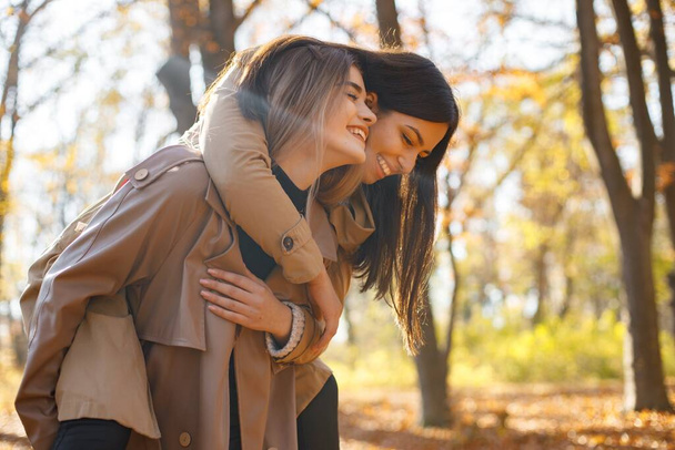 Two beautiful female friends spending time together. Two young smiling sisters walking in autumn park and hugging. Brunette and blonde girls wearing coats. - Foto, Imagen