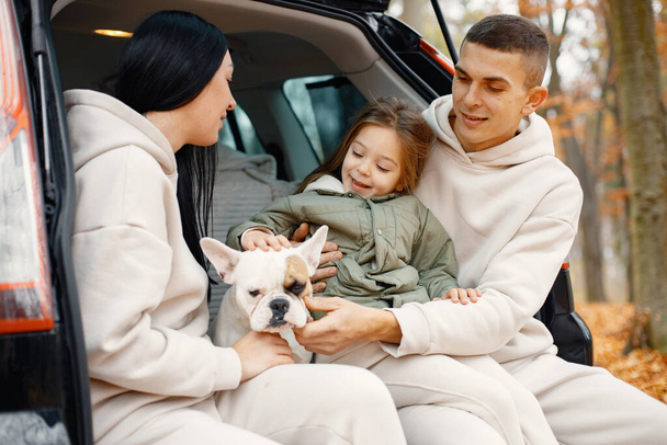 Family resting after day spending outdoor in autumn park. Father, mother, their daughter and dog sitting inside car trunk, smiling. Family wearing beige sportive costumes. - Foto, Bild