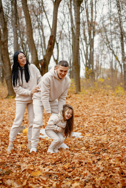 Man, woman and their little girl standing in autumn forest and laughing. Brunette woman and man play with daughter. Family wearing beige sportive costumes. - Foto, imagen