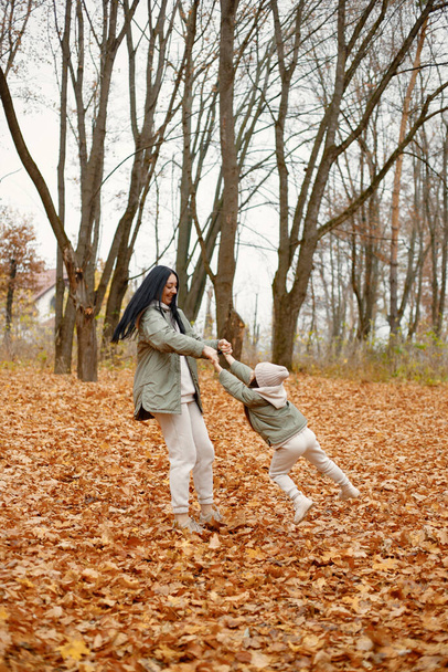女性と彼女の少女は秋の森で遊んで笑っている。ブルネットの男が娘を空中に投げ捨てた。ベージュのスポーツ衣装を着た家族. - 写真・画像