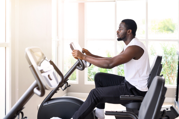 Vista lateral del hombre afroamericano haciendo ejercicio en la bicicleta estática en el gimnasio interior. Concepto saludable y aptitud física - Foto, imagen