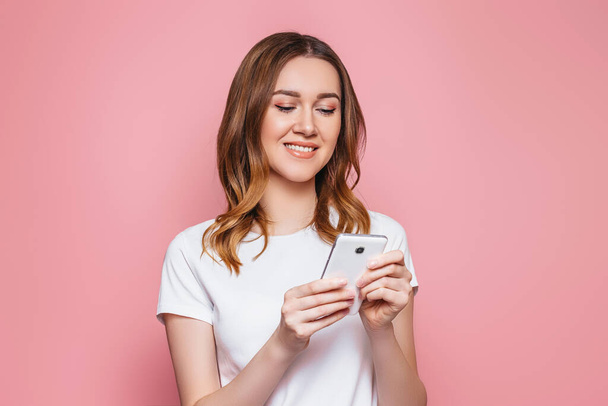 Ritratto di una giovane donna aucasica felice in una t-shirt bianca usando il telefono cellulare isolato su sfondo rosa, studentessa dai capelli ricci ondulati tiene lo smartphone e legge un messaggio, copia lo spazio - Foto, immagini