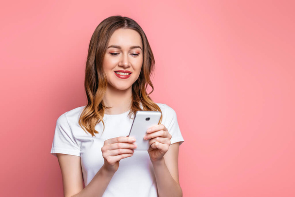 Portret van een gelukkige jonge? aucasische vrouw in een wit T-shirt met behulp van mobiele telefoon geïsoleerd over roze achtergrond, meisje student met golvend krullend haar houdt smartphone en leest een bericht, kopieer ruimte - Foto, afbeelding