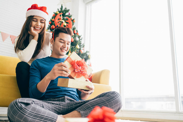 Молодая красивая счастливая азиатка в шляпе Санта-Клауса удивляет своего парня рождественским подарком дома с рождественской елкой на заднем плане. Изображение с местом для копирования. - Фото, изображение