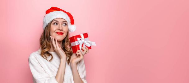 New year gifts concept. Cute caucasian young girl in a santa hat holds a box with a gift thinks and dreams makes a wish isolated on a pink background in the studio. - Photo, Image