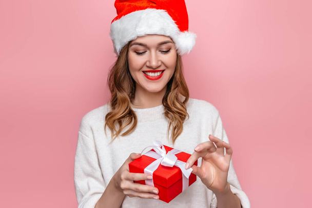Año nuevo concepto de compras en línea. Joven hermosa mujer caucásica sonriente con sombrero de santa y apertura caja de regalo roja aislada sobre fondo de estudio rosa. Copiar espacio - Foto, Imagen