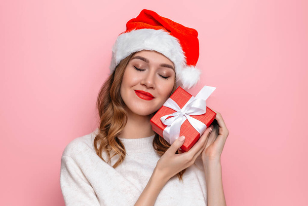 Młoda kobieta z czerwoną szminką w białym swetrze i Santa kapelusz przytula pudełko prezent odizolowany na różowym tle studio. Koncepcja prezentów noworocznych i świątecznych - Zdjęcie, obraz