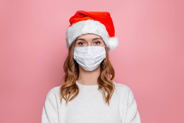 Close up portrait of sad girl in santa hat and protective medical white mask isolated on pink studio background. New Year in quarantine during the coronavirus pandemic - Fotografie, Obrázek
