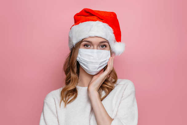 Young woman in santa hat wearing medical respiratory protective mask holds hand to face covering cheek isolated on pink studio background - Foto, Bild