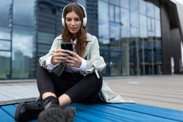 Nuori nainen kuulokkeissa, joissa on matkapuhelin, kuuntelemassa musiikkia, istuu liikekeskuksen julkisivun vieressä. - Valokuva, kuva