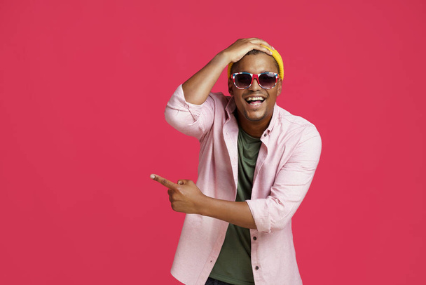 Young african american man in UK, Britain flag sunglasses, wearing casual pointing sideways excited or shocked touching his head in yellow hat isolated on red background.  - Zdjęcie, obraz