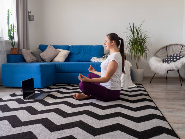 European girl looking on laptop during practising yoga. Concept of fitness, sport at home and healthy lifestyle. Online training, watching video tutorial on a laptop. Woman doing exercises on carpet. - Photo, Image