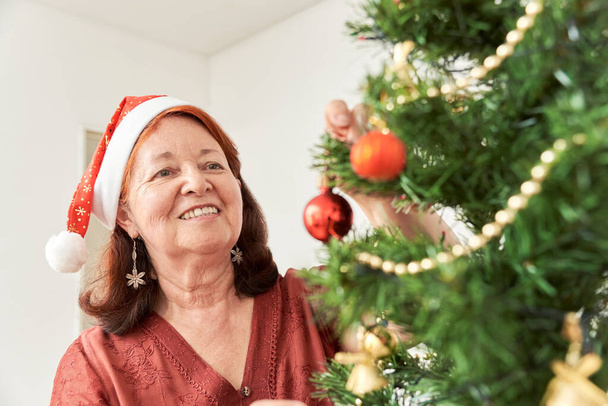 Happy senior hispanic woman smiling while decorating a Christmas tree wearing a red Santa Claus beanie. The joy of the holidays. - Photo, Image