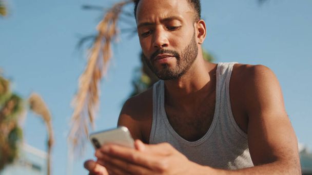 Young bearded male athlete is resting after training, sitting on bench, using smartphone  - 写真・画像