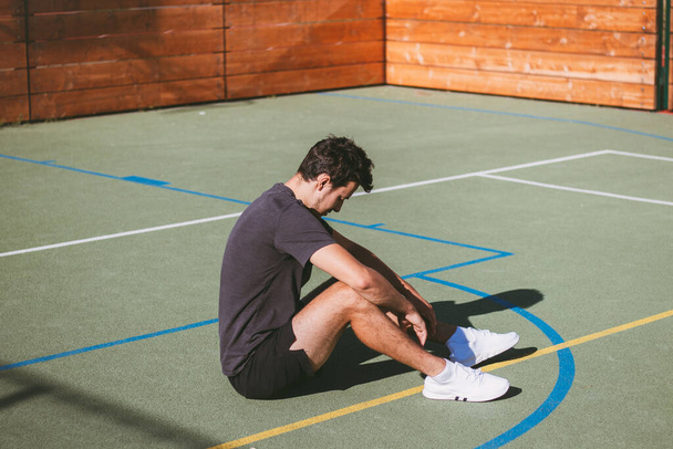 Athlète aux cheveux noirs se repose lourdement après un entraînement dur au soleil direct sur un terrain extérieur. Un athlète fatigué assis sur le sol. - Photo, image