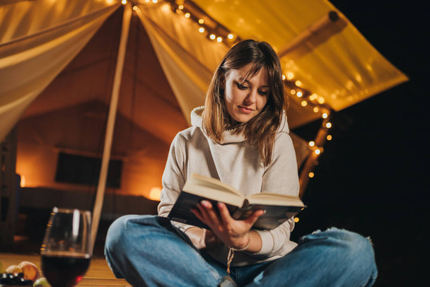 Улыбающаяся женщина-фрилансер пьет вино и читает книгу, сидя в уютной глампинговой палатке осенним вечером. Люксовый кемпинг-шатер для отдыха на свежем воздухе. Концепция образа жизни - Фото, изображение