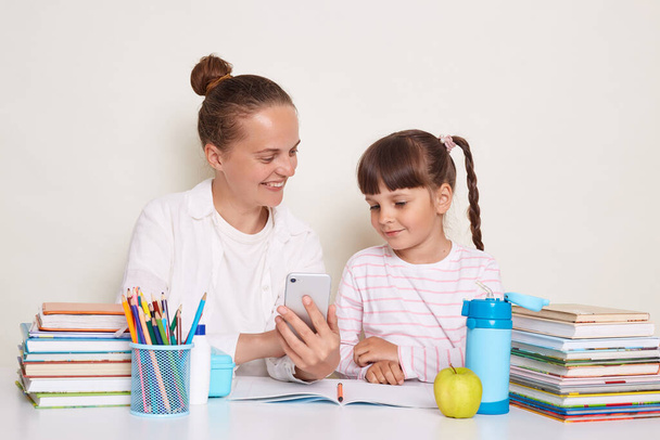 Horizontal shot of Caucasian woman with her daughter or pupil sitting at table with books doing homework, expressing positive emotions, smiling while studying, using mobile phone for doing tasks. - Photo, image