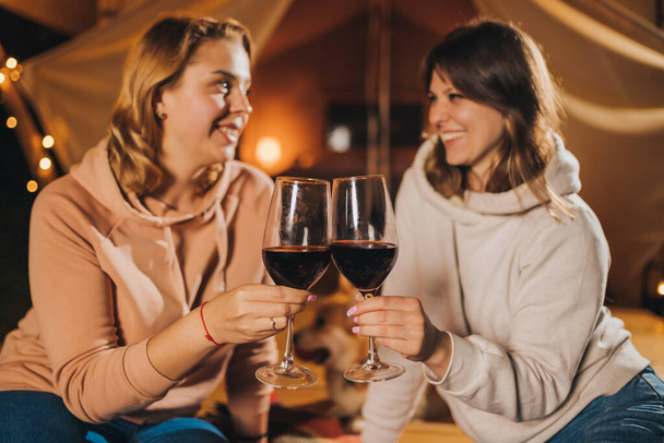 Two Smiling female friends drinking wine and eating fruits sitting in cozy glamping tent in autumn evening bonfire. Luxury camping tent for outdoor holiday and vacation. Lifestyle concept - Zdjęcie, obraz