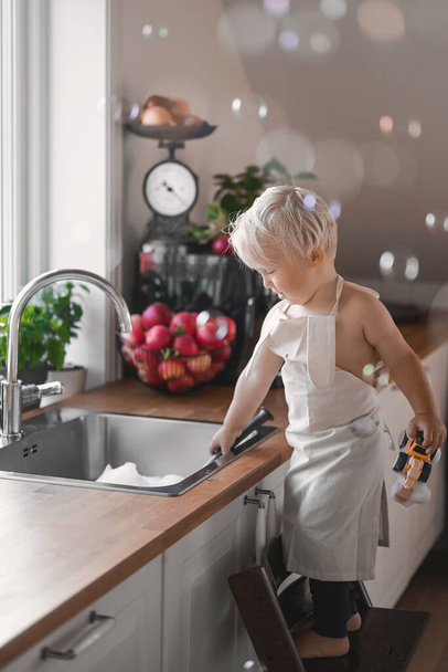 close up portrait of adorable cute baby boy washing dishes in the sink with foam and soap bubbles. - Foto, afbeelding