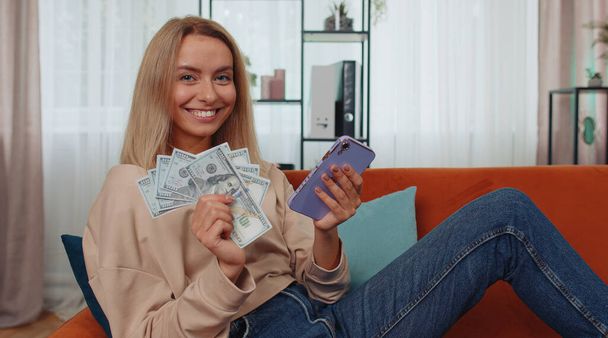 Планування сімейного бюджету. Усміхнена біла дівчина, яка рахує гроші готівкою, використовує смартфон, щоб обчислити внутрішні рахунки вдома. Радісна молода жінка задоволена доходами і заощаджує гроші на заплановану відпустку, подарунки
 - Фото, зображення