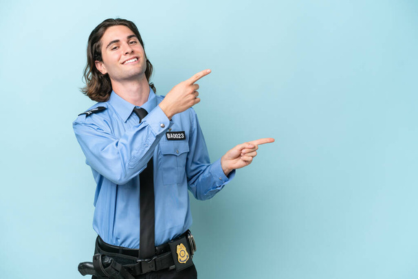 młody policjant biały człowiek odizolowany na niebieskim tle wskazując palcem w bok i prezentując produkt - Zdjęcie, obraz