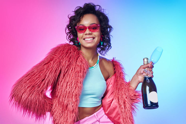 Beautiful African woman in trendy clothes holding bottle with sparkling wine against colorful background - Foto, imagen