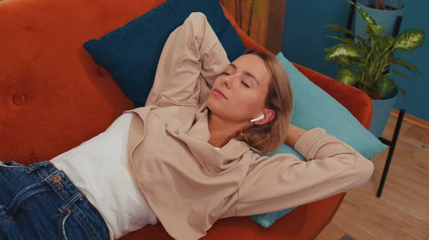 Νεαρή γυναίκα σε ασύρματα ακουστικά χαλαρώνοντας ξαπλωμένη στον καναπέ στο σπίτι επιλέγοντας ακρόαση αγαπημένο ενεργητική disco rock n roll χορευτική μουσική στο smartphone. Δραστηριότητες Σαββατοκύριακου. Χαρούμενο χαρούμενο κορίτσι - Φωτογραφία, εικόνα