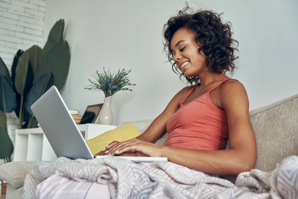 Cheerful young African woman using laptop while sitting on the couch at home - Zdjęcie, obraz