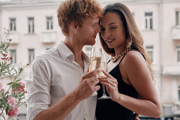 Beautiful young couple embracing and toasting with champagne while standing on the balcony together - Foto, imagen