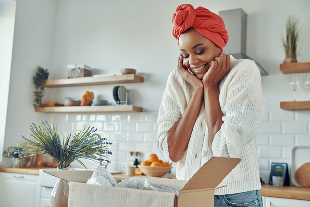 Excited African woman looking at the opened box and touching face while standing at kitchen - Foto, Bild