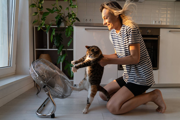Belföldi macska próbál menekülni a szőke tulajdonos utálja és ellenáll a hideg levegő áramlását kis ventilátor a konyhában. Fiatal nő megpróbál segíteni neki szeretett kisállat hűvös le a nyári hőség hangulatos lakás - Fotó, kép