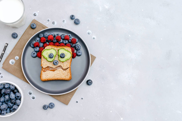 Funny scary monster face smile on halloween sandwich toast bread with peanut butter, blueberry, raspberry on plate,bats on gray background close up. Kids child sweet dessert breakfast lunch food. - Valokuva, kuva