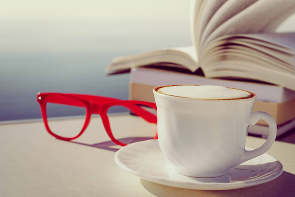Reading on holidays. Coffee cup and books on table outdoors against blue sea water background. Book pages fluttering in the wind. - Photo, image