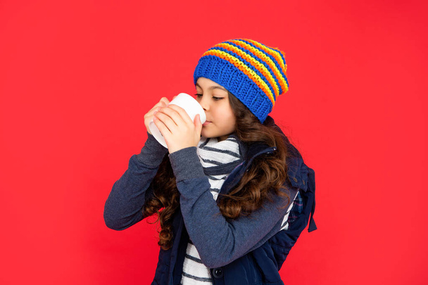 portrait of child with coffee cup. drinking cocoa and milk. kid in puffer waistcoat and hat. teen girl drink warm tea on red background. - Foto, imagen
