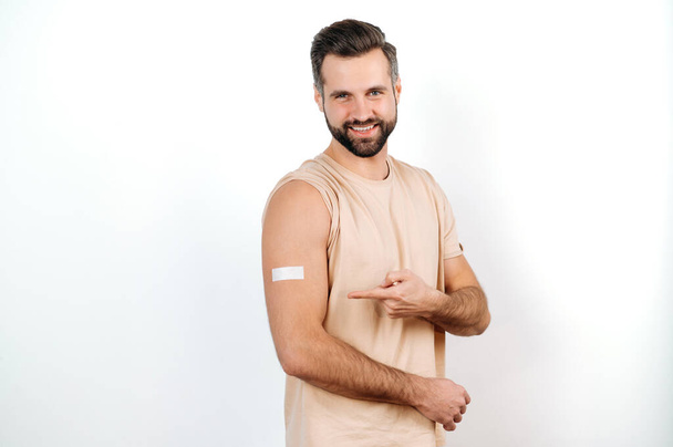 Happy caucasian man, with a plaster on his shoulder, received a vaccine, points a finger at it, stands on a white isolated background, looks at camera, smile. Vaccination, disease prevention - Фото, изображение