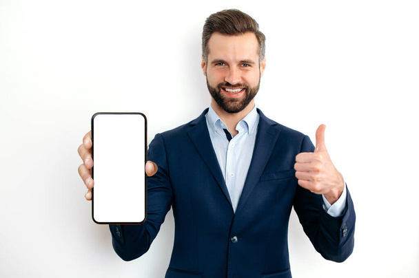 Happy confident caucasian bearded man, in a suit, showing smart phone with empty mock-up screen in hand for advertising, points finger thumb up gesture, standing on isolated white background, smiles - Foto, immagini
