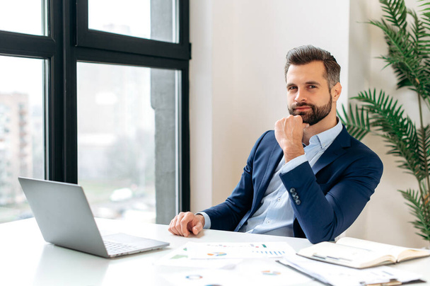 Photo of positive handsome elegant caucasian man, entrepreneur, company ceo, sitting at workplace in the office, dressed in formal suit, looking at camera. Attractive successful bearded businessman - Photo, Image