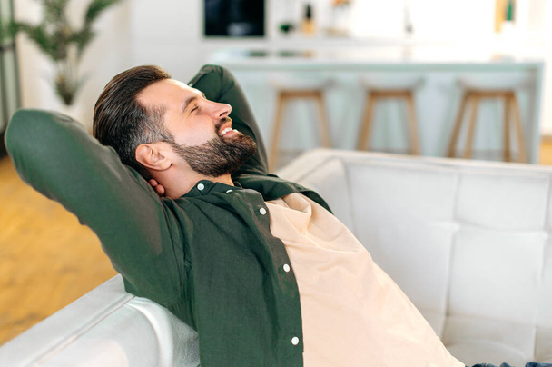 Satisfeito calmo homem barbudo caucasiano feliz, em uma camisa casual, senta-se em um sofá na sala de estar com as mãos atrás da cabeça, fechando os olhos, relaxa depois de um dia de trabalho, descansa, sorri - Foto, Imagem