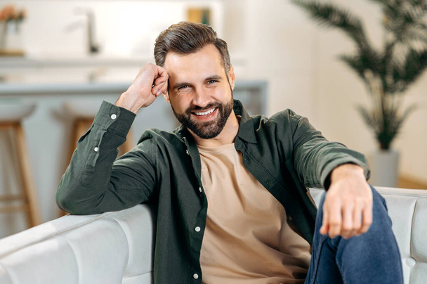 Male portrait. Headshot of a handsome attractive positive modern bearded caucasian man, in casual stylish clothes, sits on a sofa in the living room, looks at the camera, smiles friendly - Photo, Image