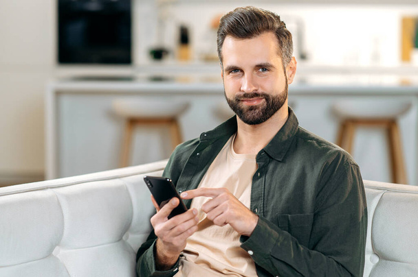Positive attractive caucasian bearded stylish man, sitting at home on a sofa in a living room, using his smart phone, relaxing on couch, messaging online in social media, looks at camera, smiles - Photo, image