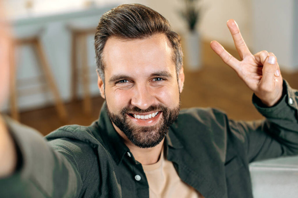 Freudig positiver kaukasischer Bärtiger im lässigen Hemd macht ein Selfie mit seinem Smartphone, zeigt mit den Fingern ein Friedenszeichen, blickt in die Kamera des Telefons, lächelt freundlich - Foto, Bild