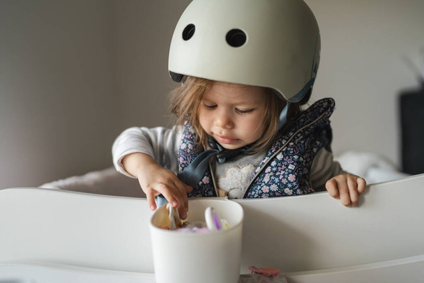 One small caucasian girl little toddler standing a t home playful with helmet on head ready to go out childhood family and growing up concept copy space - Photo, Image