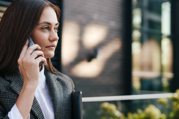 Atrakcyjna bizneswoman brunetka rozmawiająca z klientem przez telefon komórkowy, stojąca z laptopem przed biurem. Odbiór. Biznesowy sukces. Telefon komórkowy - Zdjęcie, obraz