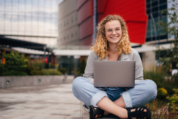 Vooraanzicht van een glimlachend studentmeisje dat brillen draagt met behulp van een laptop met afstandsonderwijs zittend buiten de campus. E-Learning. Virtueel - Foto, afbeelding