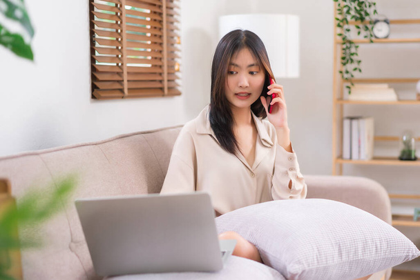 Käsite toiminnan olohuoneessa, nuori aasialainen nainen käyttää kannettavaa tietokonetta ja puhuu älypuhelimella sohvalla. - Valokuva, kuva