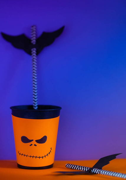 Halloween-Dekor. Papierbecher mit Stroh innen und Papiersilhouetten von Fledermäusen, heller Hintergrund. Gespenstische Feiertagssymbole, selektiver Fokus. Raum für Text - Foto, Bild
