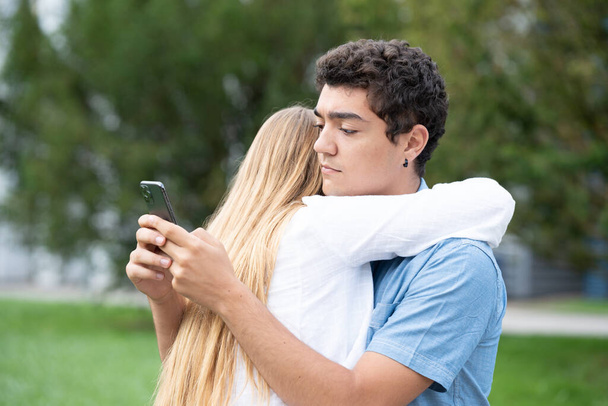 Spaanse tiener jongen knuffelen vriendin en chatten op de telefoon. Bedriegen en ontrouw concept - Foto, afbeelding
