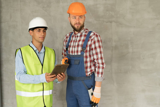 Construction worker and engineer on site discussing blueprints on pad or tablet computer, gray wall background. Place for text - Zdjęcie, obraz