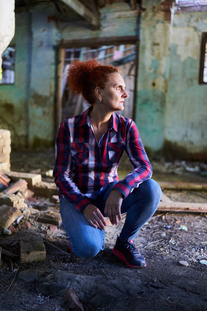 Curly haired redhead woman in plaid shirt and blue jeans in a ruined barn and house - Foto, Bild
